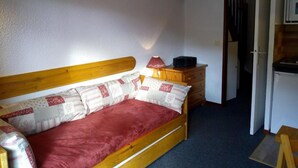 Una piccola camera con un letto singolo, una testiera in legno, un materasso rosso e cuscini decorati. Si vede un comò in legno con una lampada, un tavolino e una porta che conduce a un'altra stanza.