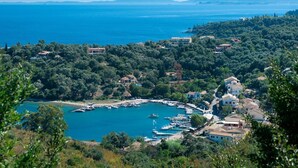 Un village côtier dont les maisons sont entourées d’une végétation luxuriante, un port de plaisance avec des bateaux, et une vue dégagée sur la mer et les montagnes lointaines.