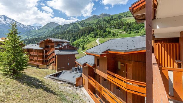 Ein Chalet-Stil-Gebäude aus Holz mit mehreren Balkonen vor einer grünen Hügel- und Berglandschaft.