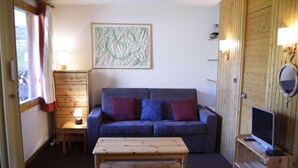 Une pièce chaleureuse avec un plancher en bois, un canapé bleu agrémenté de coussins rouges et bleus, une table basse en bois, une lampe, un téléviseur posé sur un meuble en bois et un miroir fixé au mur.