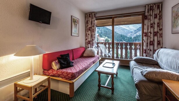 A hotel room with a red sofa, a grey sofa, a small table, a TV mounted on the wall, and a balcony with a mountain view. - 90m from ski slopes, 4th floor, view mountain, balcony, 36m², Méribel-Mottaret (Méribel - Les Allues)