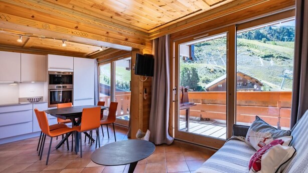 A modern interior with a wooden ceiling, a dining area with orange chairs, a sofa with patterned cushions, and large windows offering a view of a mountainous landscape. - 130m from ski slopes, 1st floor, view resort, balcony, ski locker, 44m², Méribel-Mottaret (Méribel - Les Allues)