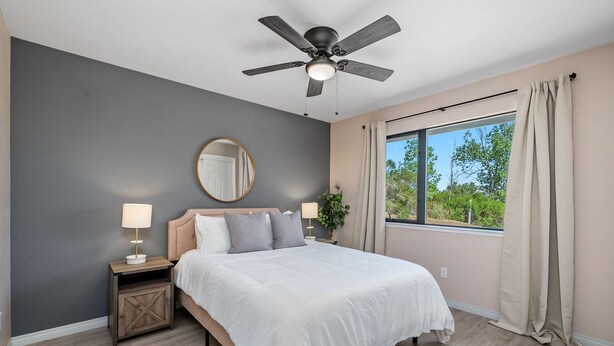 A bedroom with a bed, a ceiling fan, a window with curtains, a bedside table with a lamp, and a mirror on the wall. - Pool Table + Firepit + BBQ | Eastside Paso Wine Country | Casa Merlot (Paso Robles)
