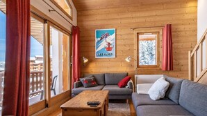 Ein gemütliches Wohnzimmer mit Holzboden, einem grauen Sofa, einem Holztisch und einem großen Fenster mit roten Vorhängen.