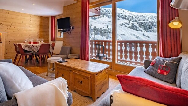 Ein gemütliches Zimmer mit Holzvertäfelung, einem Essbereich mit Tisch für vier Personen und einem großen Fenster mit Blick auf verschneite Berge.