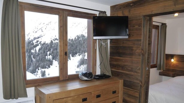 Ein Schlafzimmer mit einem Holzkleiderschrank, einem an der Wand montierten Fernseher, einem Bett mit weißer Bettwäsche und Blick auf eine verschneite Berglandschaft durch ein großes Fenster.
