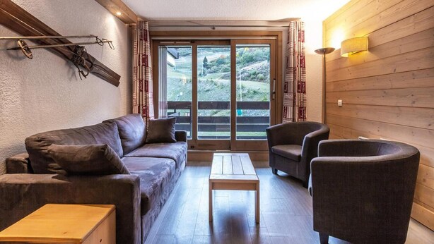A cozy living room with a sofa, armchair, and coffee table, a wooden wall, and a large window with curtains. - 80m from ski lifts, 4th floor, balcony, ski locker, 46m², Méribel-Mottaret (Méribel - Les Allues)