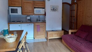 Eine kleine Küche mit Holzausstattung, einer Mikrowelle, einer Kaffeemaschine und einem Spülbecken. Es gibt einen Esstisch mit Stühlen und ein violettes Sofa.