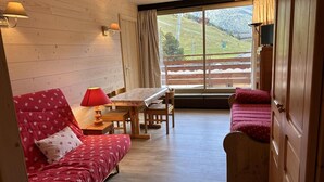 A cozy room with a red patterned sofa, a wooden dining table with chairs, and a view of a snowy landscape through a large window. - 20m from ski slopes, 3rd floor, view mountain, balcony, ski locker, 24m², Méribel-Mottaret (Méribel - Les Allues)