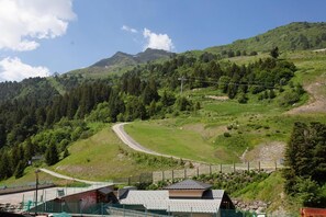 Ländliche Berglandschaft mit einer kurvenreichen Straße, einem kleinen Gebäude mit Satteldach und einem eingezäunten Bereich mit grünem Netz.