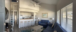 Relax in this stylish living area with a sofa, coffee table, and bar area, featuring a ceiling fan and a window with blinds.