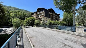 Un puente que lleva a un edificio de varios pisos con balcones, rodeado de vegetación y colinas.