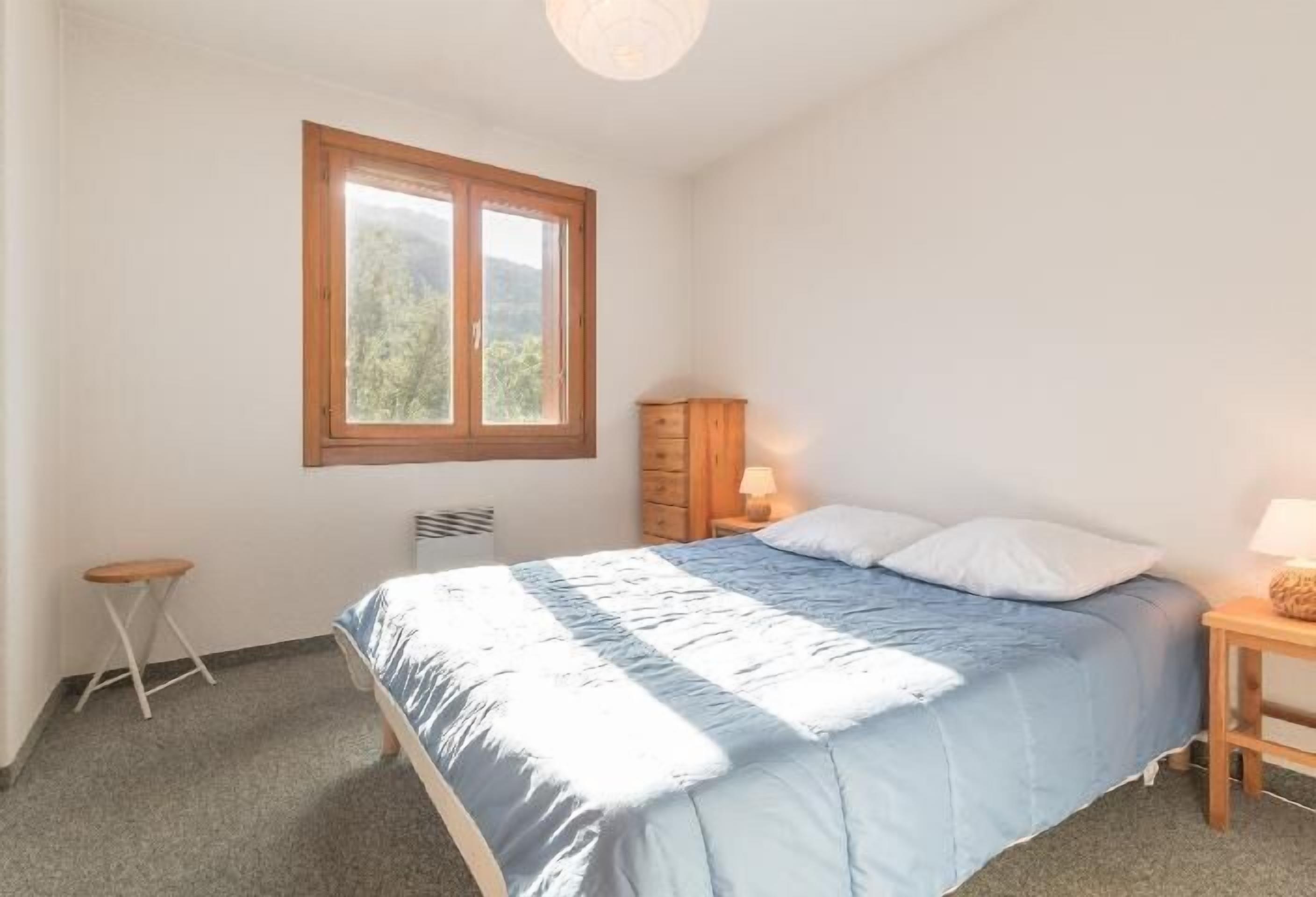 A bedroom with a bed, a window, a nightstand, a lamp, and a small stool.