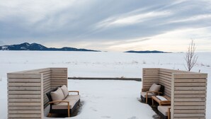 A snow-covered outdoor seating area with wooden benches and a cushioned bench, surrounded by a snowy landscape and distant mountains. - Luxury Stand Alone Condo, Minutes from Downtown Bozeman, Garage & Outdoor Patio (Bozeman)