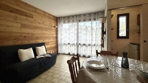 A room with a wooden wall, a dark sofa, a dining table with chairs, and a window with sheer curtains. - 350m from ski slopes, 3rd floor, view mountain, tv, 25m², Serre Chevalier (Briançon)