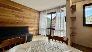 A room with a wooden wall, a black sofa, a dining table with a white tablecloth, and a window with a view of a building and trees. - 350m from ski slopes, 3rd floor, view mountain, tv, 25m², Serre Chevalier (Briançon)