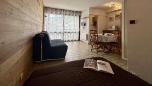 A cozy room with a wooden wall, a dark sofa, a dining area with a table and chairs, and a kitchen area with a microwave. - 350m from ski slopes, 3rd floor, view mountain, tv, 25m², Serre Chevalier (Briançon)