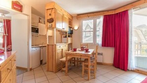 A compact kitchen with wooden cabinets and a dining table, a red curtain, and a view of a house through the window.