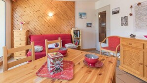 A cozy room with a wooden ceiling, a red sofa, a wooden dining table with a bowl and a box, and a bookshelf.
