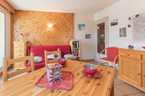 A cozy room with a wooden ceiling, a red sofa, a wooden dining table with a bowl and a box, and a bookshelf. - 200m from resort center, 3rd floor, view mountain, balcony, tv, 33m², Serre Chevalier (Briançon)