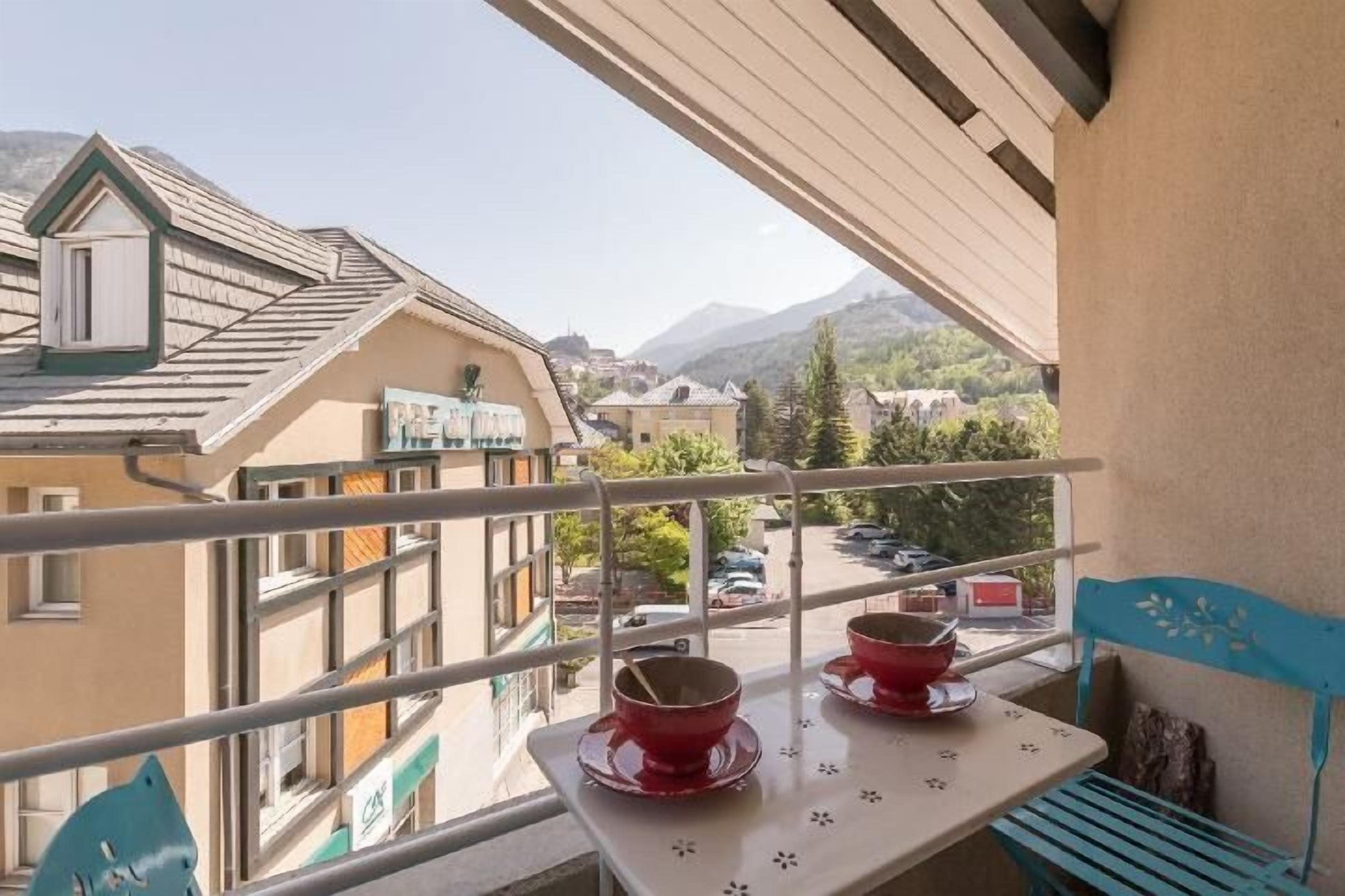 A balcony with two red bowls on a table, a blue chair, and a view of buildings and mountains in the background.