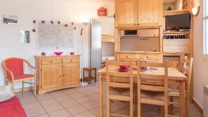 A compact kitchen with wooden cabinets, a dining table with chairs, a red chair, and a small stool.