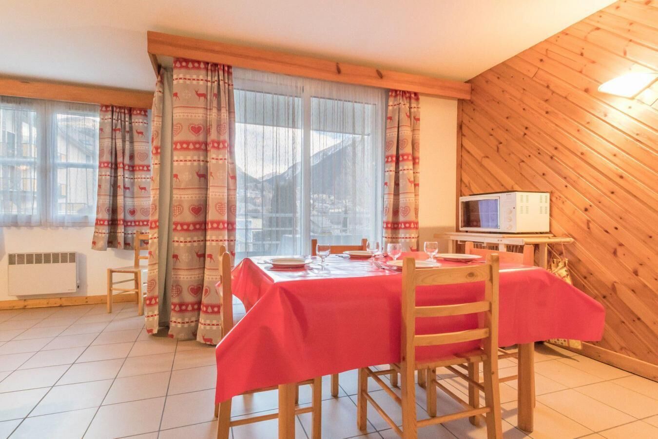A dining area with a table set for four, wooden chairs, a red tablecloth, and a television on a wooden stand.