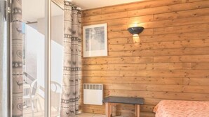 Une pièce aux murs recouverts de lambris en bois, un lit avec une couvre-lit à motifs floraux, un banc, une lampe fixée au mur, un tableau encadré et une porte-fenêtre coulissante avec des rideaux à motifs.