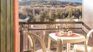 Un balcon avec une table et des chaises, donnant sur un paysage vallonné parsemé d’arbres et offrant une vue lointaine sur un télésiège.