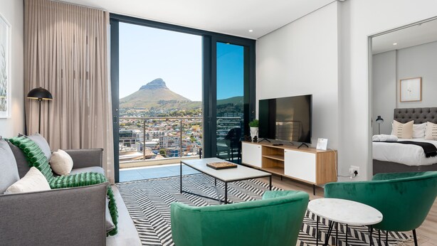 Ein modernes Hotelzimmer mit großem Balkon, einer Couch, einem grünen Sessel, einem kleinen Tisch und einem Fernseher.