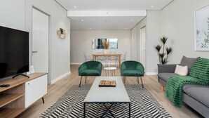 Ein modernes Wohnzimmer mit Fernseher, einer Couch, grünen Sesseln, einem Couchtisch und einem Essbereich mit Tisch und Stühlen.