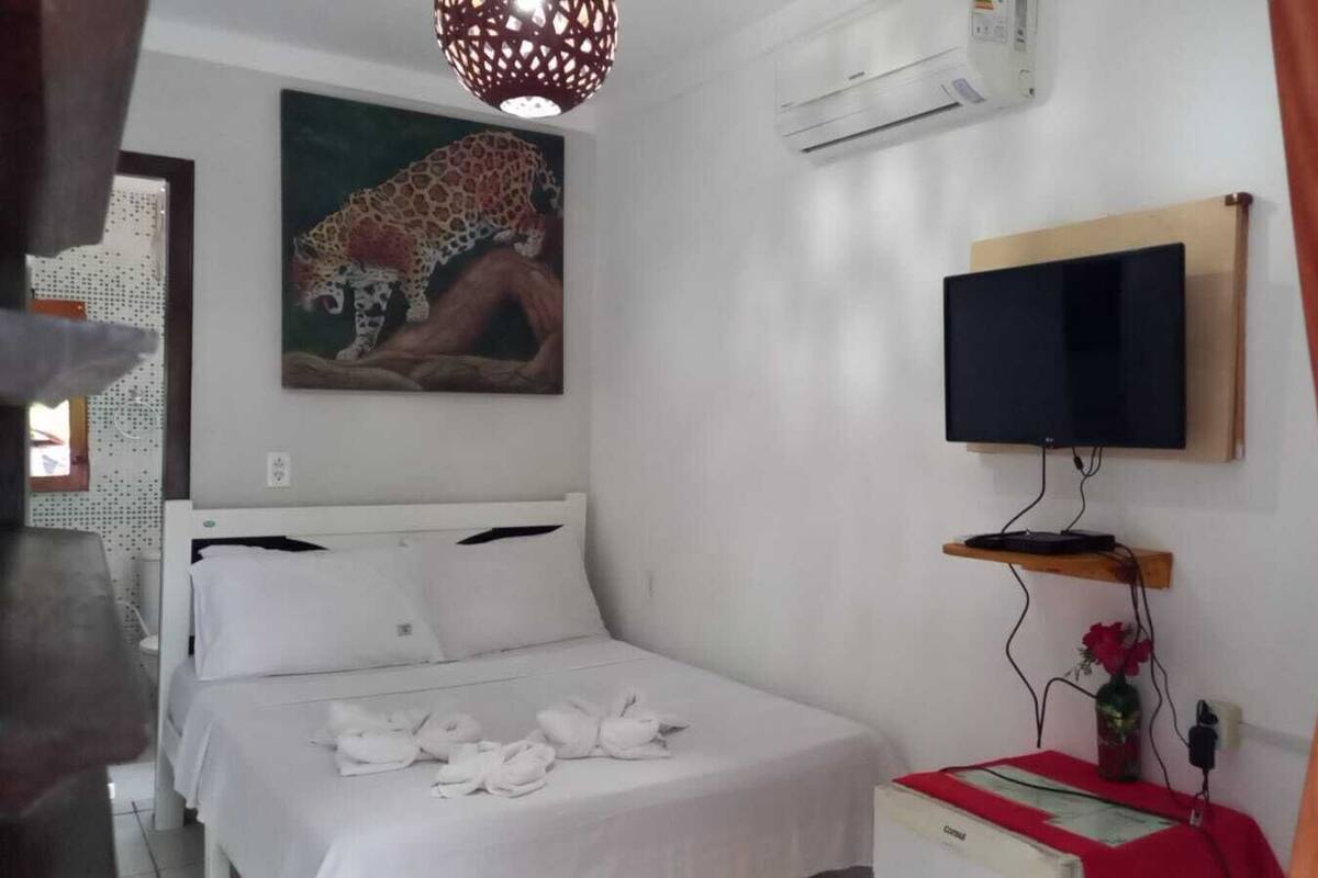 A bedroom with a bed, a TV mounted on the wall, a painting, and an air conditioning unit.