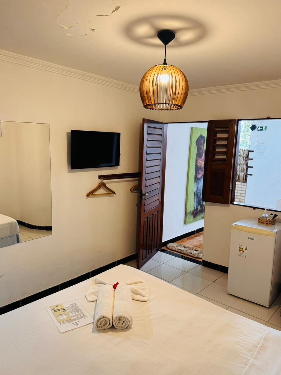 Quarto de hotel com cama, televisão, pequeno frigorífico e candeeiro suspenso.
