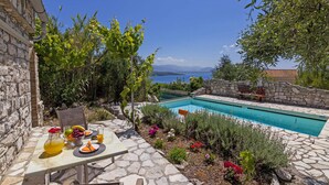 En pool av sten med fri utsikt över havet och bergen, omgiven av frodig grönska och en stenmur.