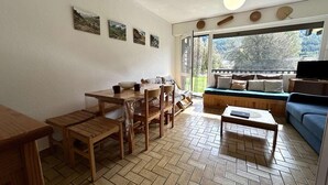 A living room with a wooden dining table, chairs, a sofa, and a coffee table. There are four framed pictures on the wall and a large window providing a view of the outdoors. - Terrace, balcony, parking, 28m², Serre Chevalier (Le Monêtier-les-Bains)