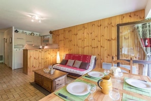 A cozy room with a wooden wall, a red sofa, a wooden dining table set for two, and a kitchen area with cabinets and a refrigerator.