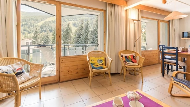 Ein gemütliches Zimmer mit Korbmöbeln, einem Esstisch und großen Fenstern mit Blick auf einen bewaldeten Berg.
