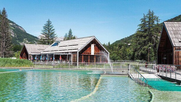 A rustic wooden building with a swimming pool in front, surrounded by mountains and trees.