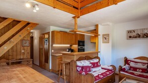 A cozy cabin interior with a wooden dining table, two cushioned chairs, a kitchen area with a bar, and a loft bedroom with a staircase. - 175m from shops, 3rd floor, view mountain, balcony, tv, 60m², Méribel-Mottaret (Méribel - Les Allues)