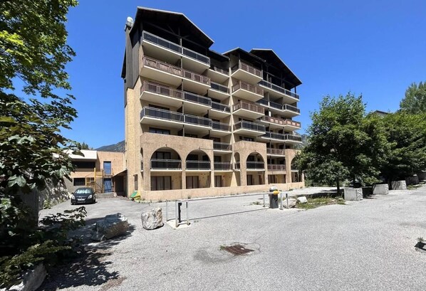 A multi-story hotel with balconies, a parking area, and surrounding greenery.