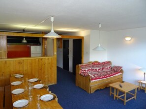 A room with a dining table set for four, a sofa, a small table with a remote, and a kitchen area in the background.