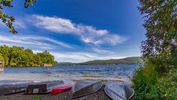 A lakeside view with several boats docked on the shore, surrounded by lush greenery and a clear sky. - Lake George 5 Bed/ 3 Bath home w/ lake & resort access (Lake George)