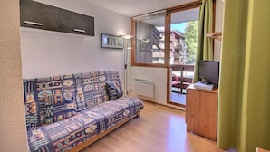 Un petit salon avec un canapé à motifs, un téléviseur posé sur un meuble en bois et une porte-fenêtre coulissante donnant sur un espace extérieur.