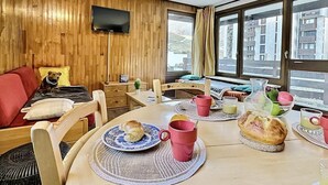 Une pièce chaleureuse avec un mur en bois, un téléviseur fixé au-dessus d’un lit, une table à manger avec des aliments pour le petit-déjeuner et une fenêtre donnant sur la ville.