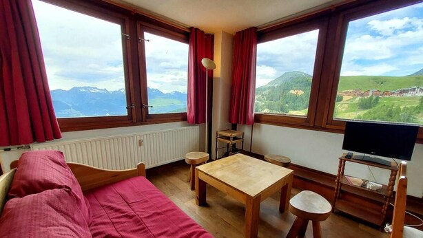 Habitación con cama, una mesa de madera, un mueble para televisión y ventanas amplias con vistas a la montaña.