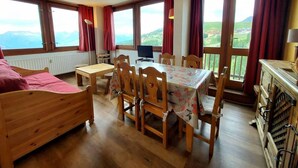 Una habitación con suelo de madera, una cama, una mesa de comedor con sillas, un televisor y ventanas amplias con cortinas.