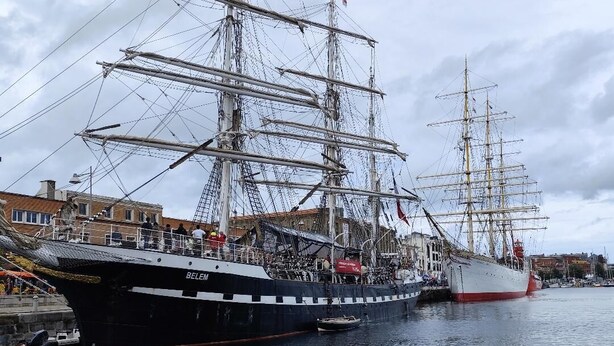 Een groot zeilschip afgemeerd in een haven, met een stadsgezicht op de achtergrond.