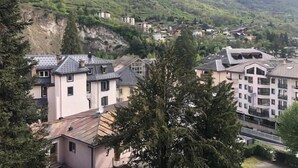A hillside residential area with houses, some with sloped roofs and others with flat roofs, surrounded by greenery and mountains in the background.