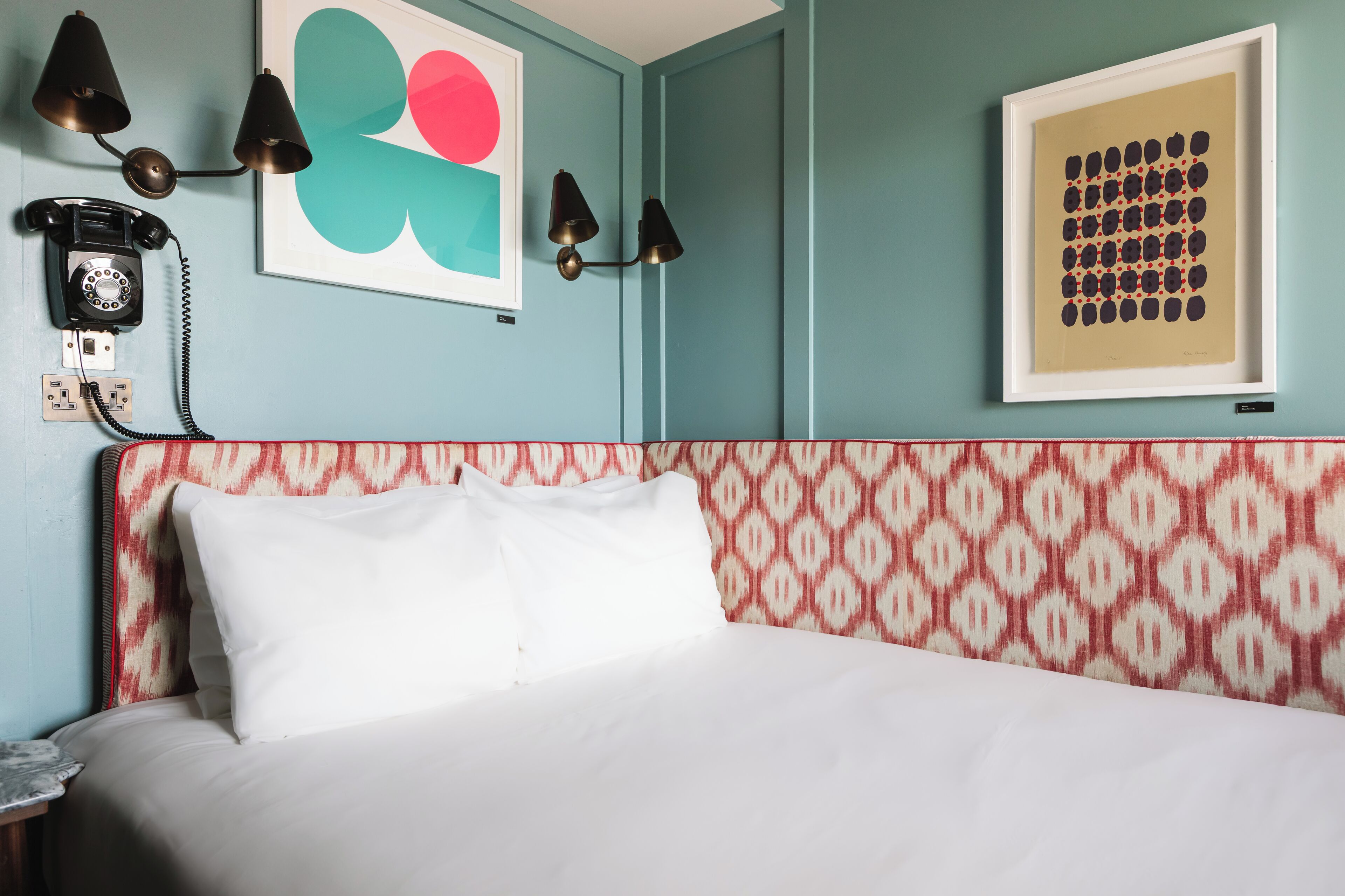 A hotel room with a bed, a telephone, wall art, and a patterned headboard.