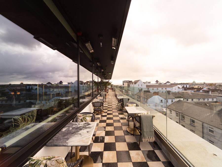 A balcony with a checkered floor, tables, and chairs, overlooking a cityscape with buildings and a cloudy sky.